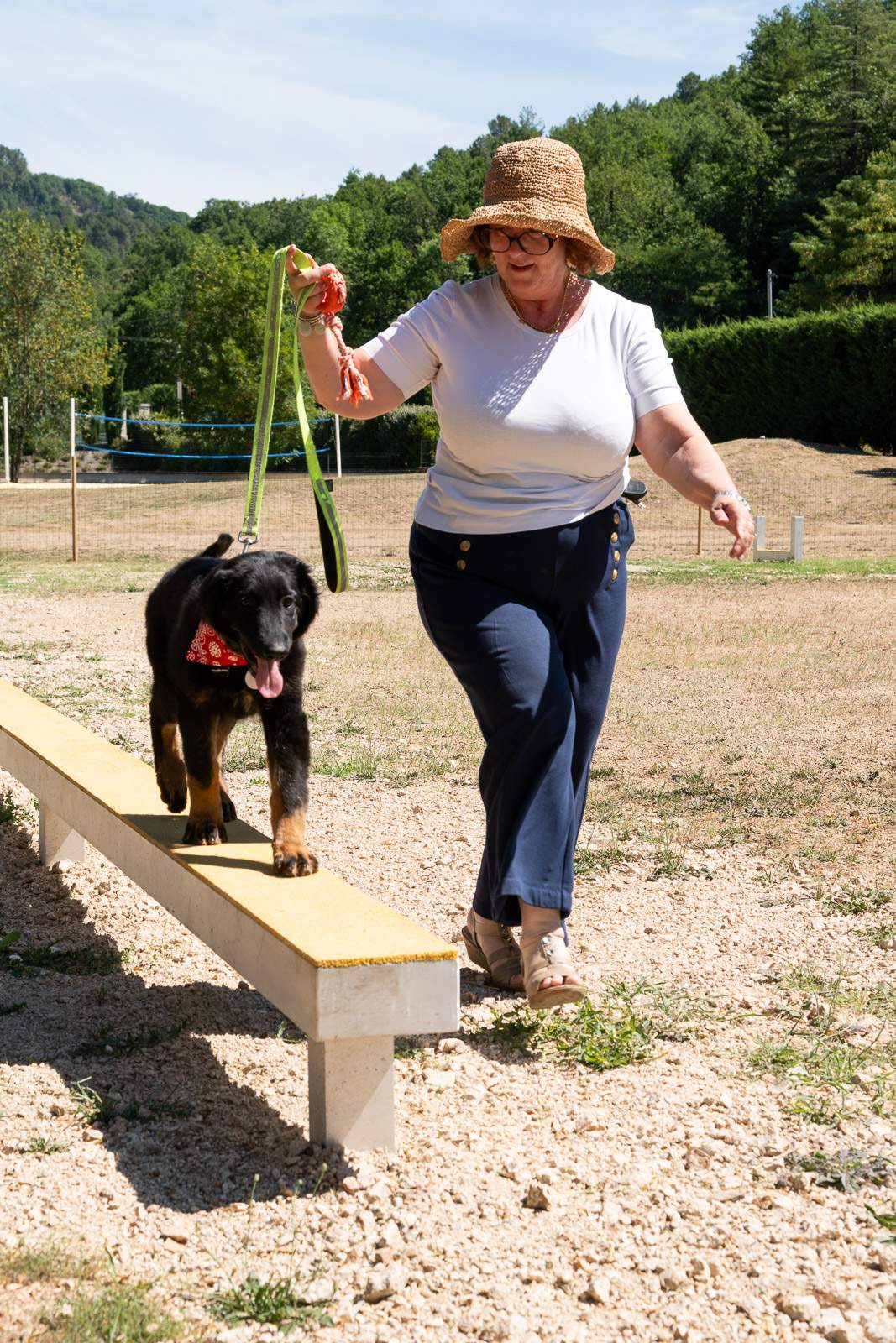 Une femme âgée portant un chapeau de paille et des lunettes marche sur une aire en plein air en tenant une laisse verte, tandis qu’un chien noir à collier rouge court près d’elle sur une petite rampe de pierre.