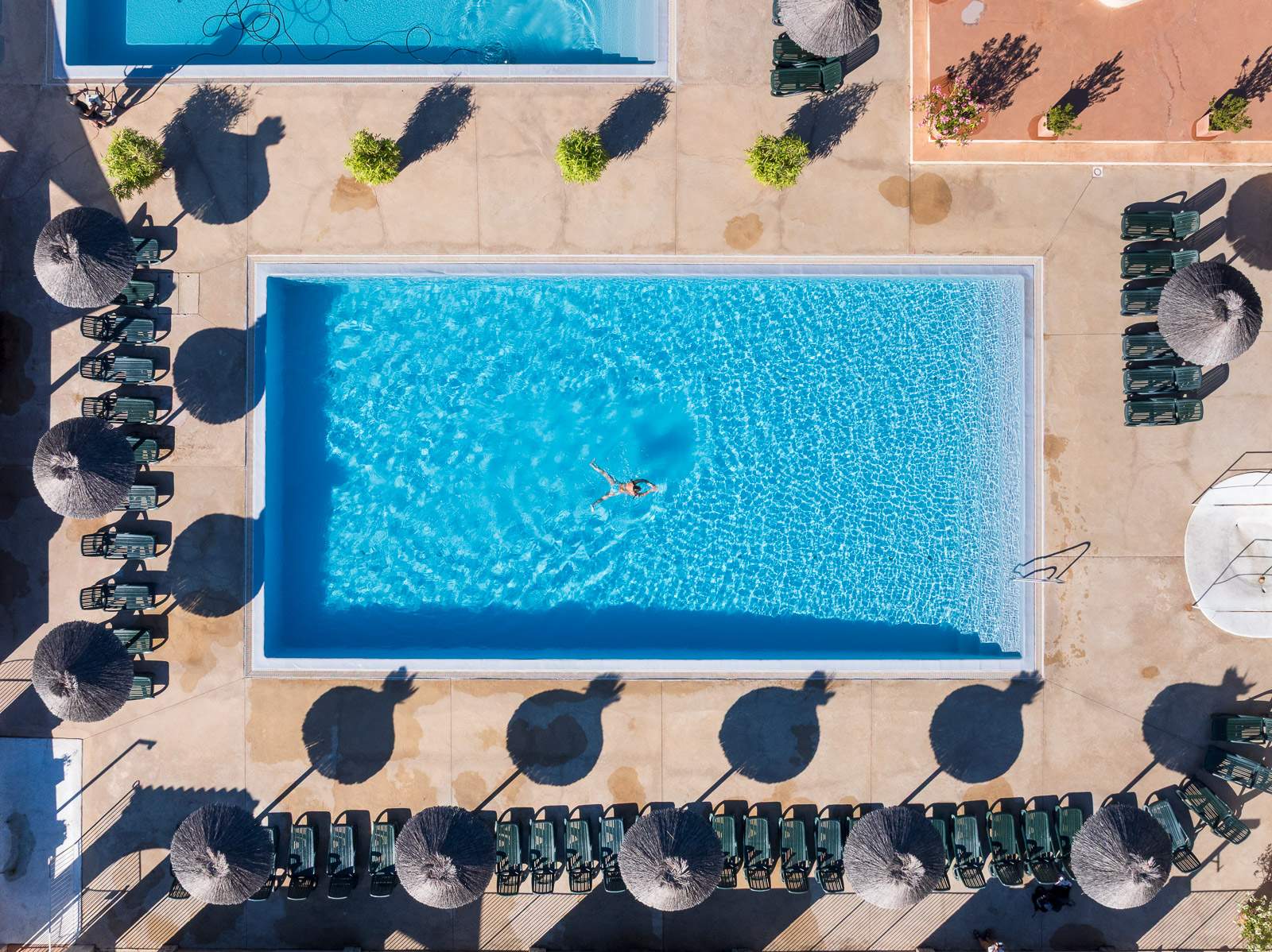 Vue aérienne d’une piscine rectangulaire remplie d’eau bleue, entourée de chaises longues et de l’ombre de parasols, avec un nageur au centre et des plantes sur les bords.