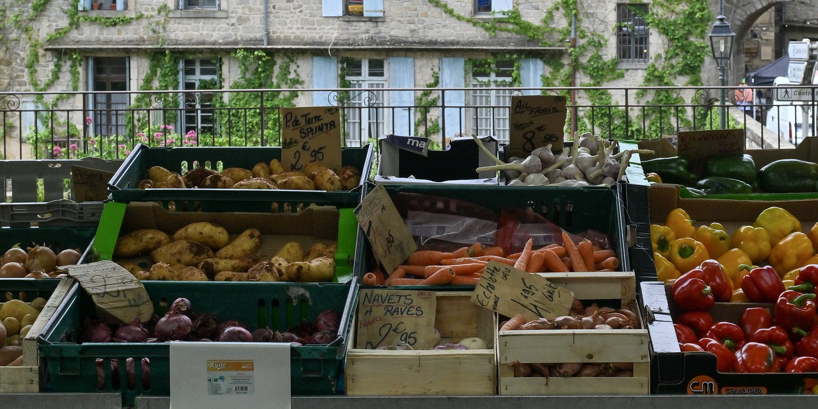 Cageots de pommes de terre, carottes, oignons et poivrons avec leurs ardoises de prix sur un étal du marché de Largentière