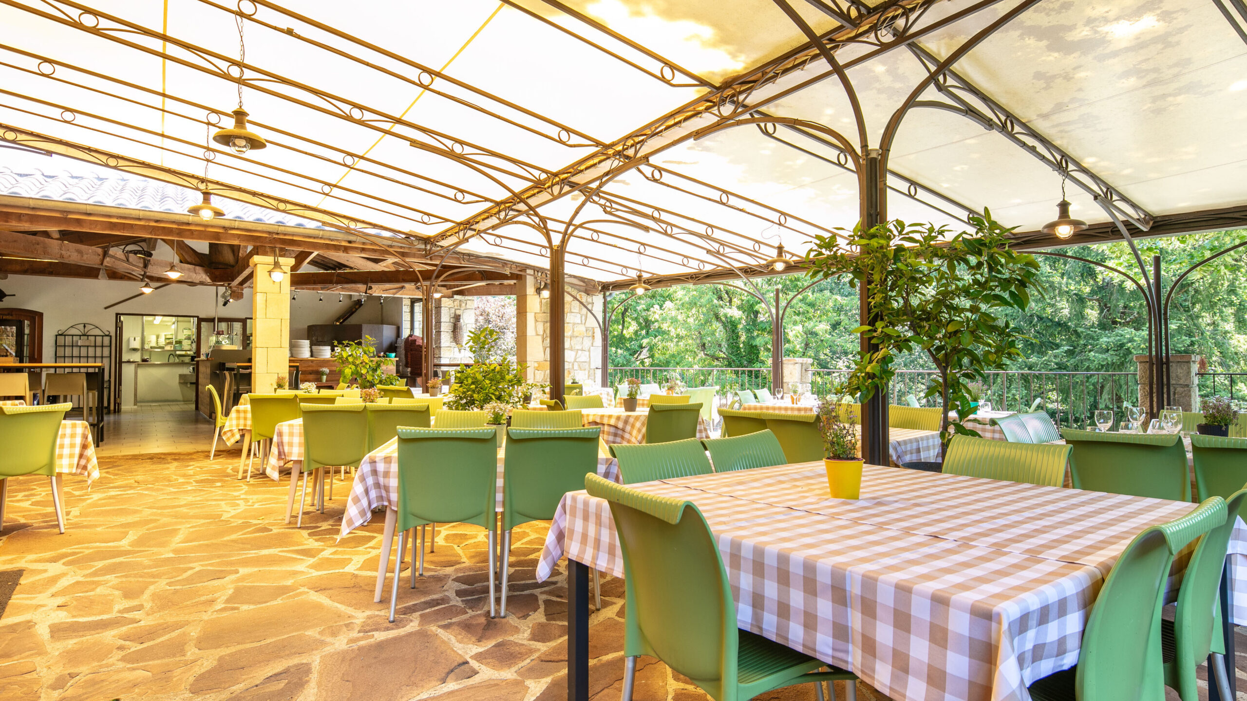 Terrasse couverte d'un restaurant avec tables nappes à carreaux vichy, chaises vertes, structure métallique Art nouveau et vue sur jardin verdoyant.