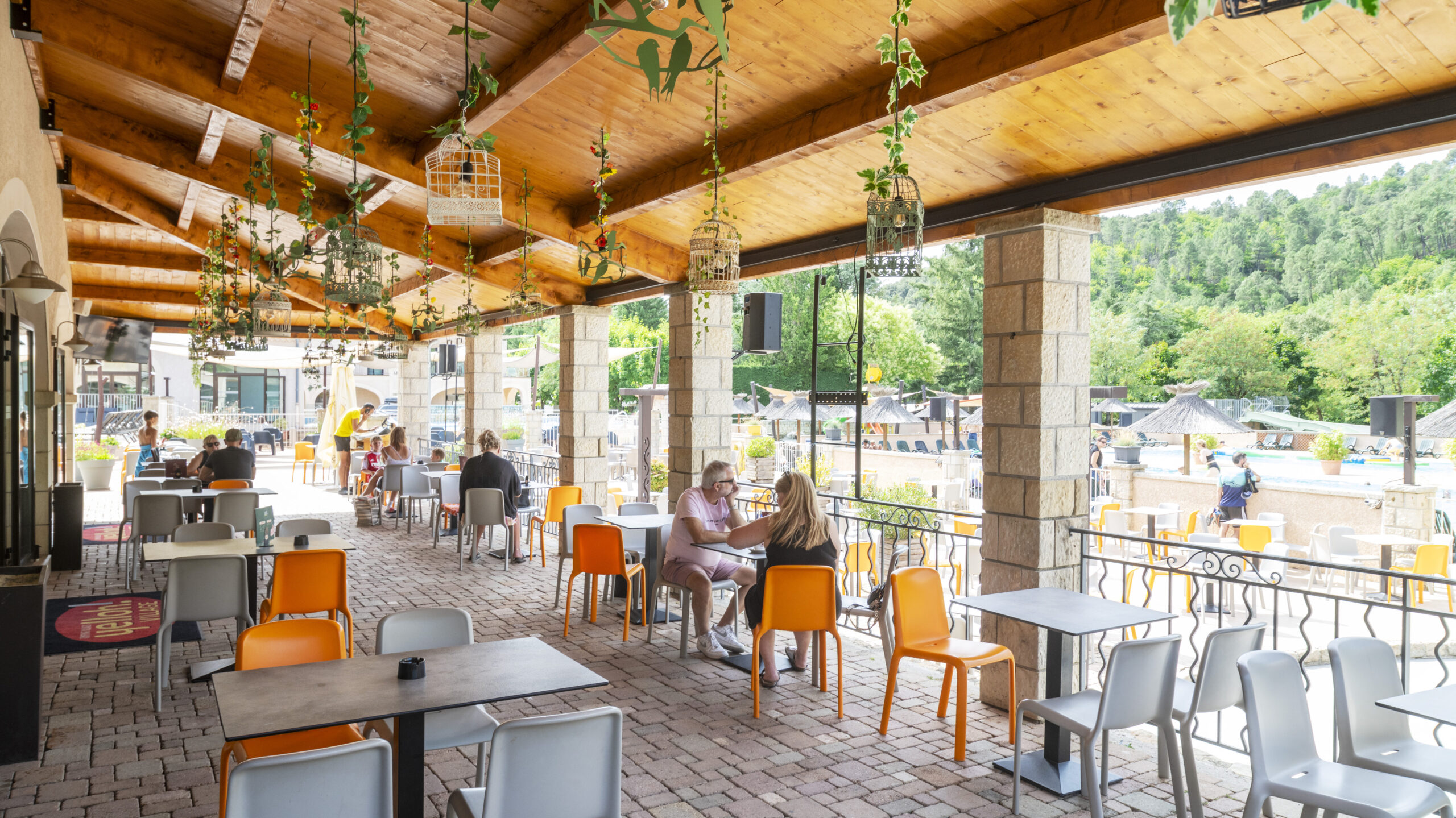 Terrasse couverte d'un restaurant avec plafond en bois apparent, plantes suspendues, chaises oranges et grises, vue sur une piscine et des arbres verdoyants en arrière-plan.