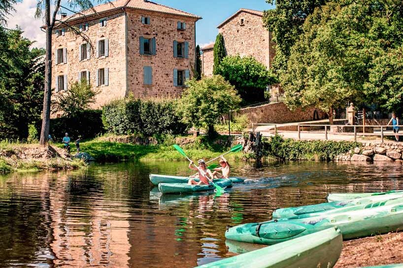 Plan d’un canal bordé de verdure avec des kayaks turquoise sur l’eau et deux personnes pagayant devant un bâtiment en pierre avec tour ronde, sous un ciel bleu.