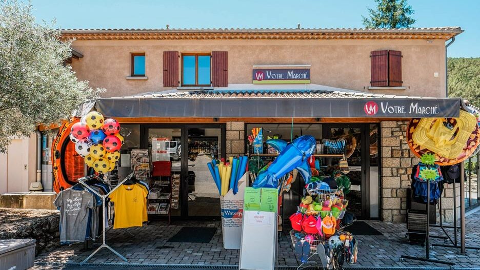 Petit magasin Votre Marché avec devanture ouverte présentant articles de plage colorés, parasols, bouées et vêtements sous un bâtiment méditerranéen ocre aux volets marron