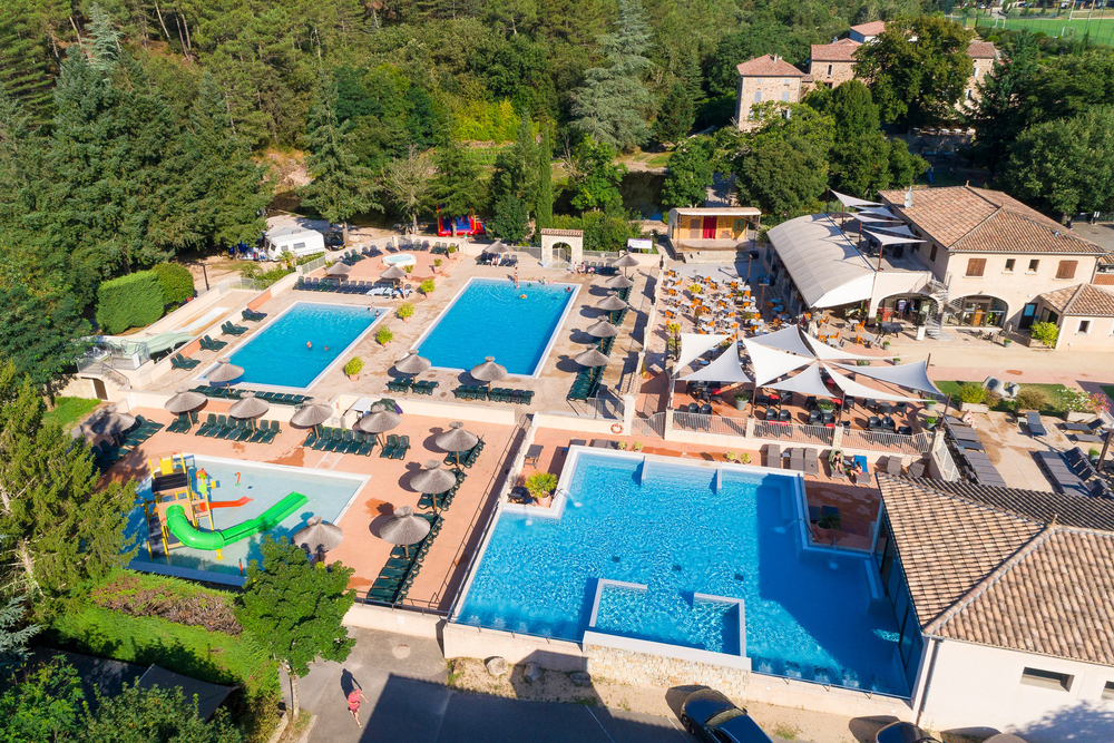 Vue aérienne d’un centre de vacances avec deux grandes piscines bleues, un bassin chauffé pour enfants, des transats et parasols sur des terrasses, et plusieurs bâtiments en pierre entourés de verdure.