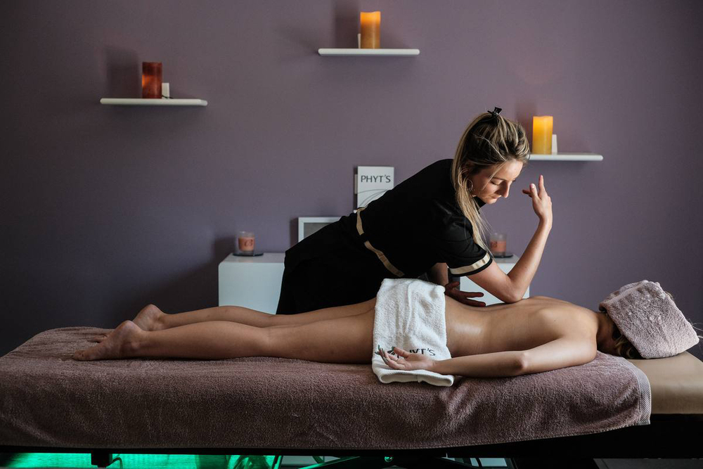 Une praticienne en massage utilise ses mains pour masser le dos d’une personne allongée sur une table de massage, dans une salle au décor apaisant avec bougies allumées.